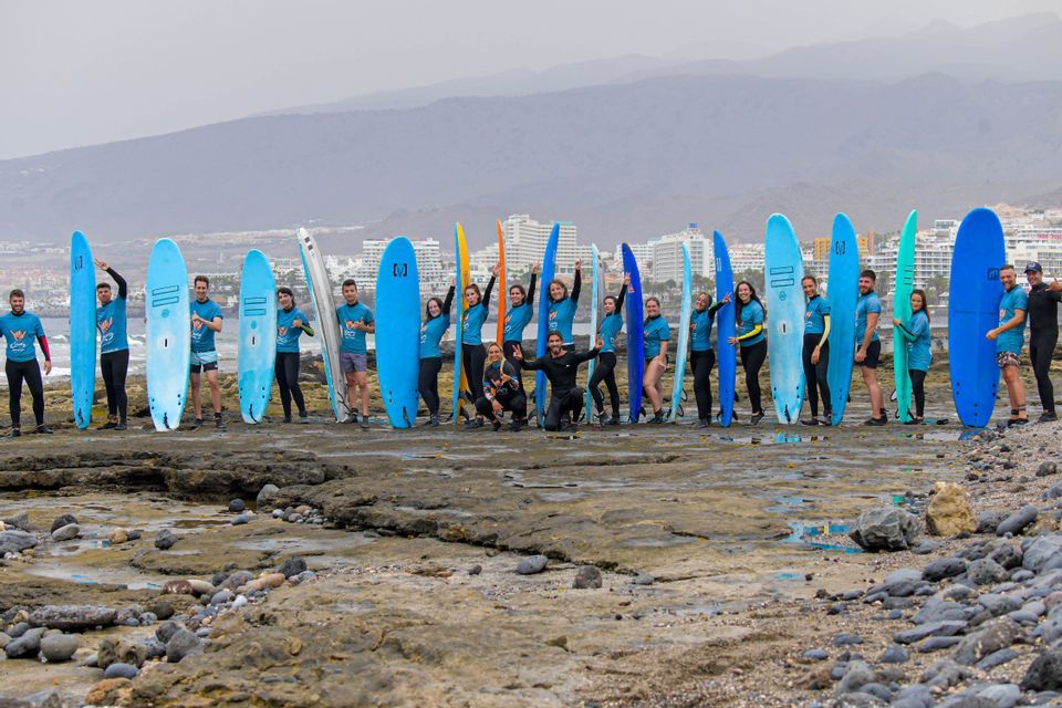Un gruppo WeRoad in posa con le loro tavole da surf colorate su una spiaggia rocciosa di fronte a una città costiera.