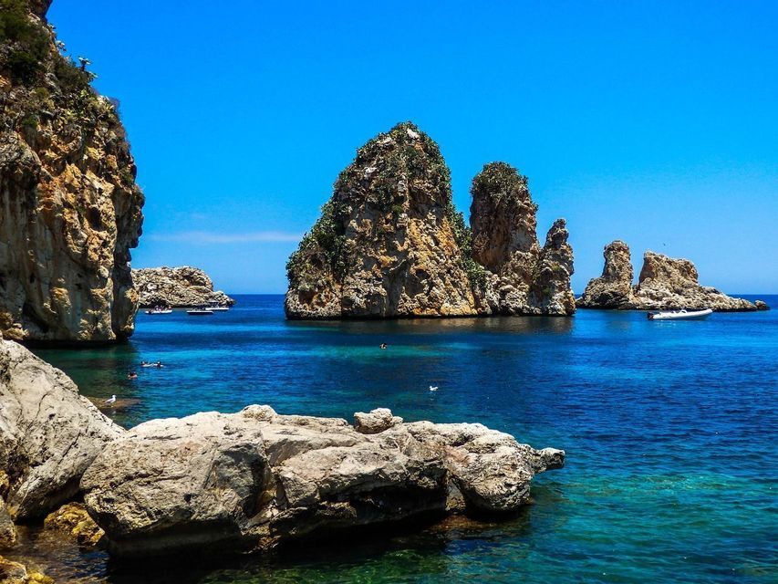 Farallones rocosos con vegetación verde emergen del agua clara y turquesa, con pequeñas embarcaciones y nadadores cerca en un día soleado.