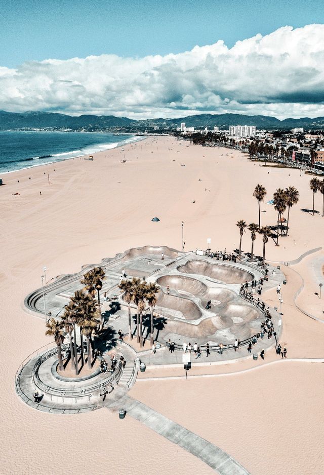 Una vista aérea de un skatepark de hormigón en una amplia playa de arena, con palmeras, el océano y montañas al fondo.