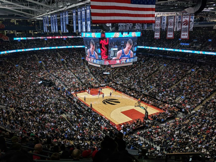 Vista aérea de un partido de baloncesto en progreso dentro de una arena abarrotada, con un gran jumbotron mostrando a un jugador.