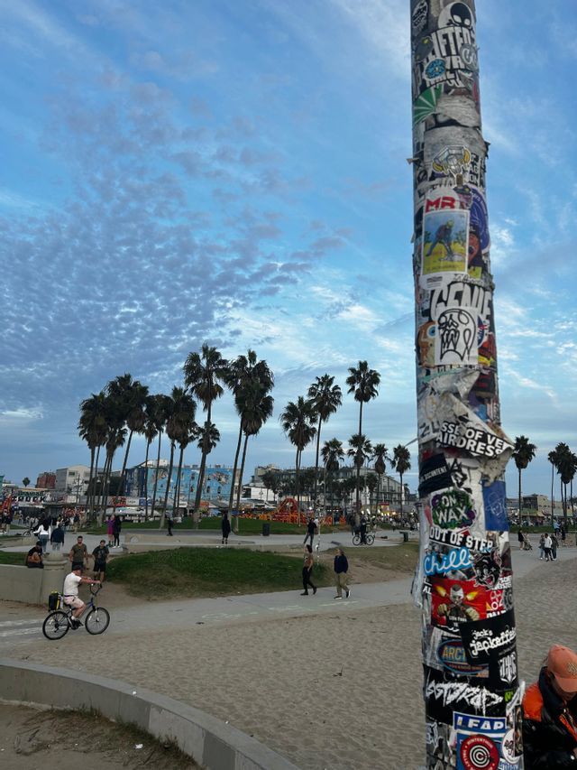 Un poste cubierto de pegatinas se alza en una playa, con gente caminando y palmeras bajo un cielo nublado al fondo.