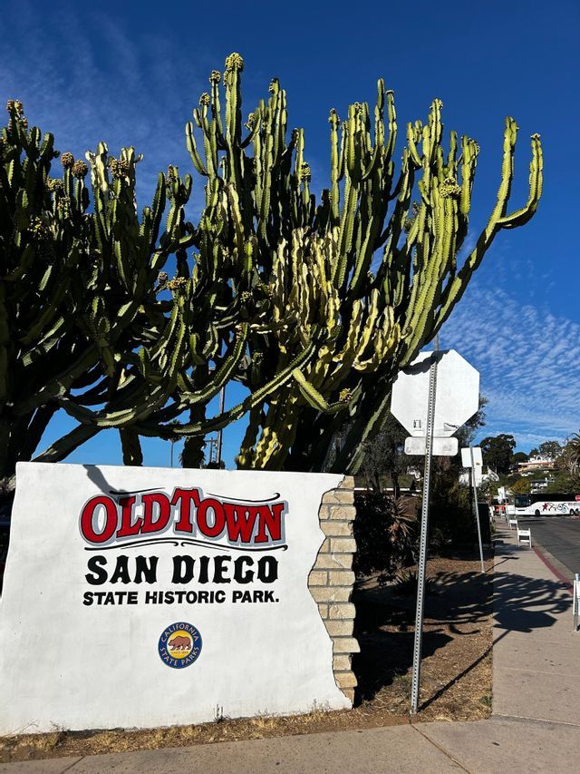 El letrero blanco de entrada del Parque Histórico Estatal Old Town San Diego se alza frente a un gran cactus bajo un cielo azul.