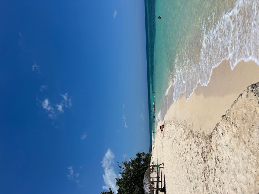 Una vista aérea de una playa de arena blanca con gente nadando en el agua turquesa bajo un cielo azul claro.