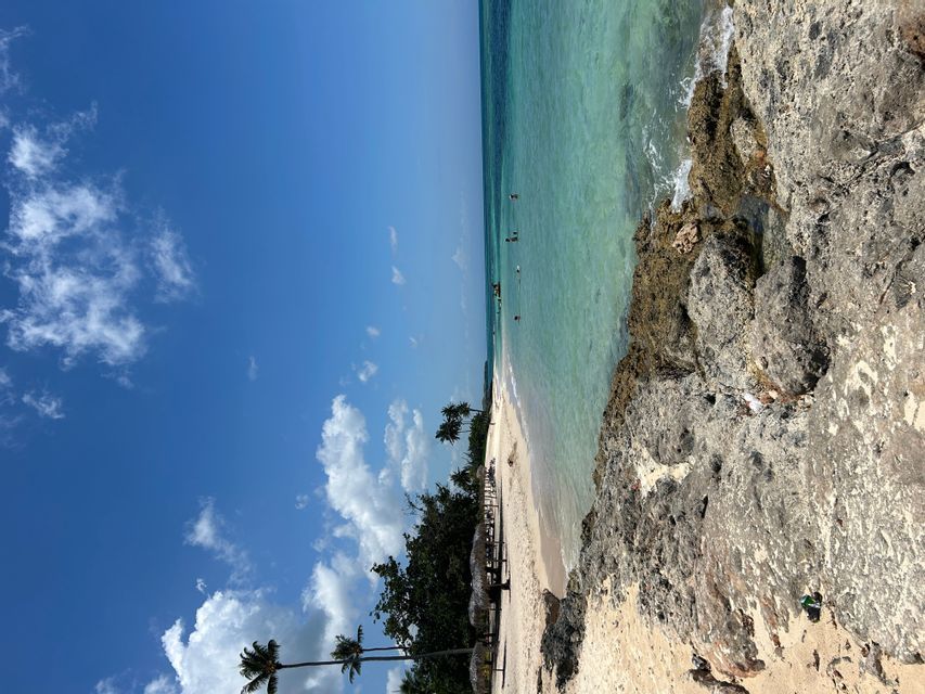 Una costa rocosa junto a una playa de arena con palmeras, agua cristalina de color turquesa y un cielo azul con nubes blancas.