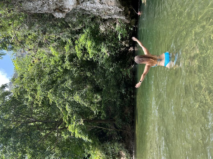 Una mujer con trenzas largas nada en un río claro y verde al pie de un bosque exuberante y un acantilado rocoso.