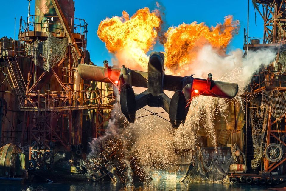 Ein Wasserflugzeug fliegt bei einer Stuntshow tief über dem Wasser, mit einer großen feurigen Explosion dahinter.