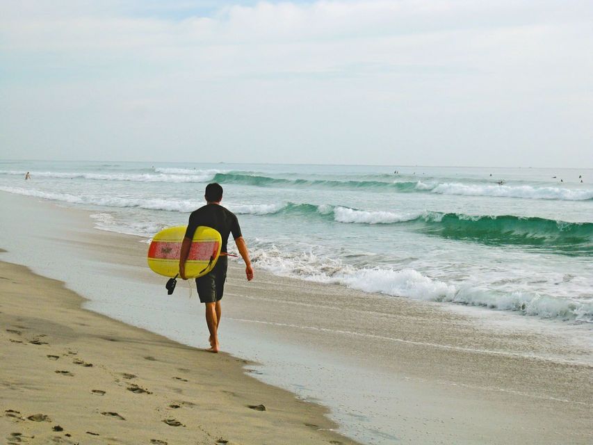 Un surfista in muta cammina lungo una spiaggia sabbiosa, trasportando una tavola da surf gialla verso il mare.