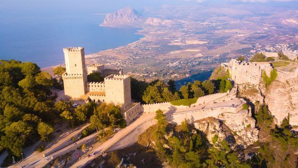 An aerial view of a stone castle with towers on a wooded mountain, overlooking a coastline with a town and the sea.
