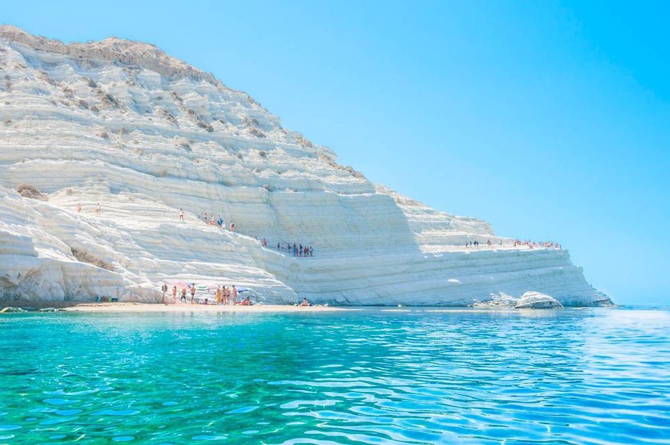 People relax on a beach and walk on large, white layered cliffs next to bright turquoise water on a sunny day.