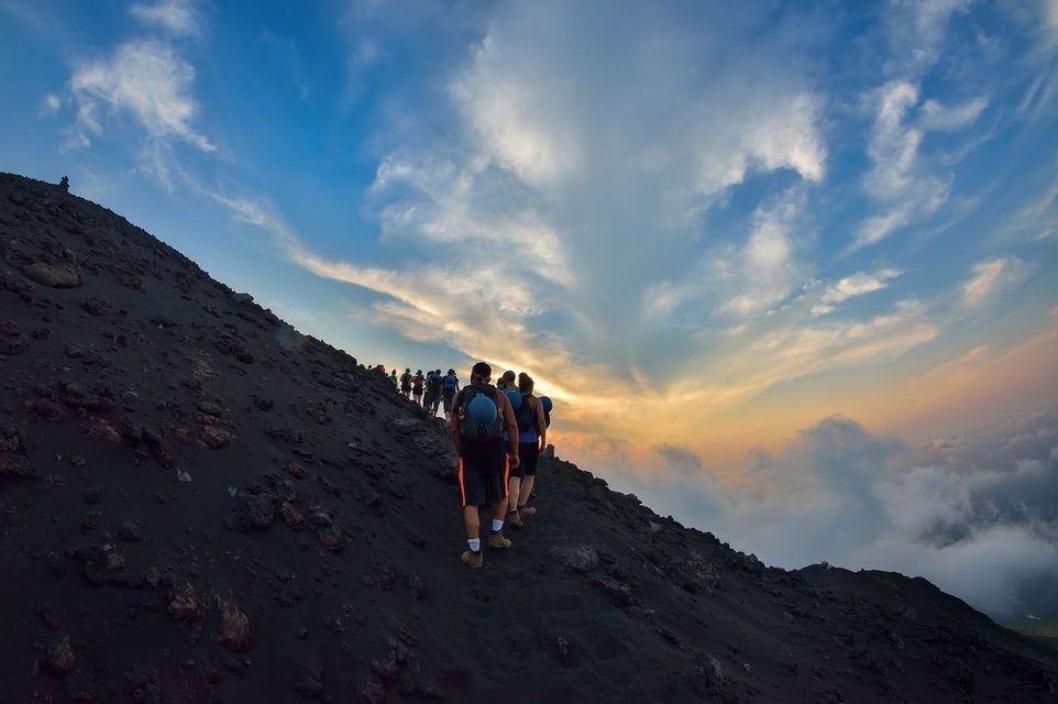 A WeRoad group trip with backpacks hiking up a steep volcanic slope under a cloudy sky at sunset.