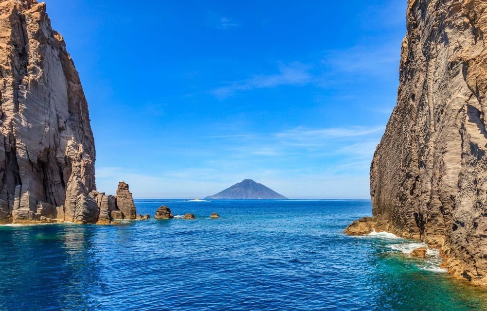 A view through two large rock cliffs of a distant conical island on a calm blue sea under a clear sky.