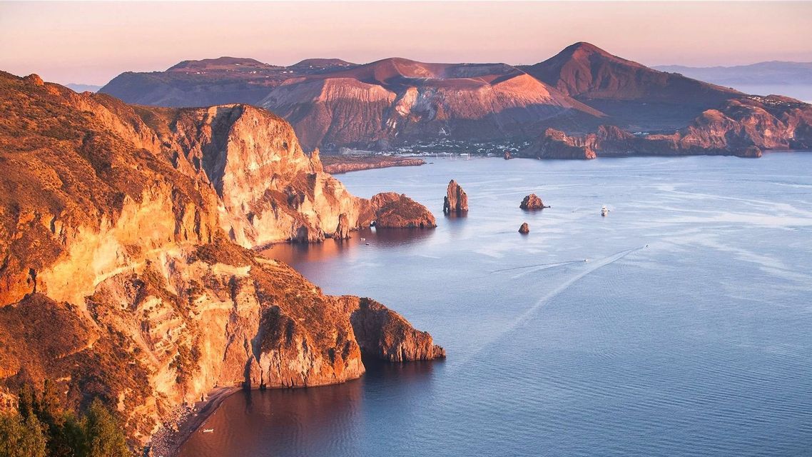 Sunlit cliffs and sea stacks overlook a calm sea with a volcanic island in the background during sunrise.