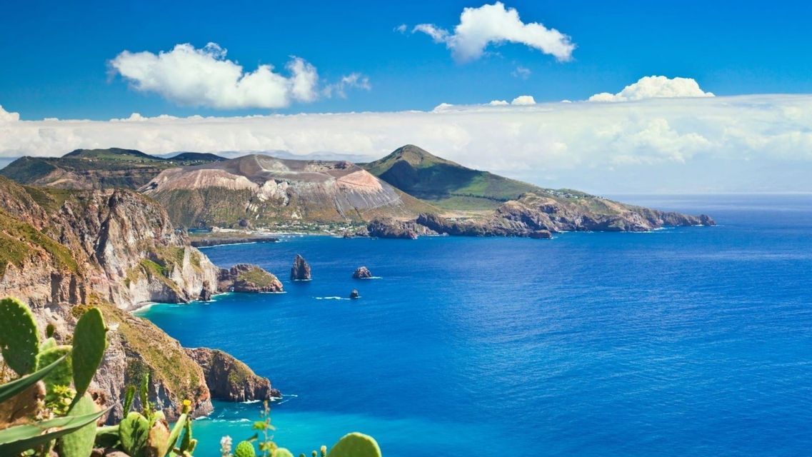 A panoramic view of a volcanic island's coastline meeting a deep blue sea, under a bright blue sky with white clouds.