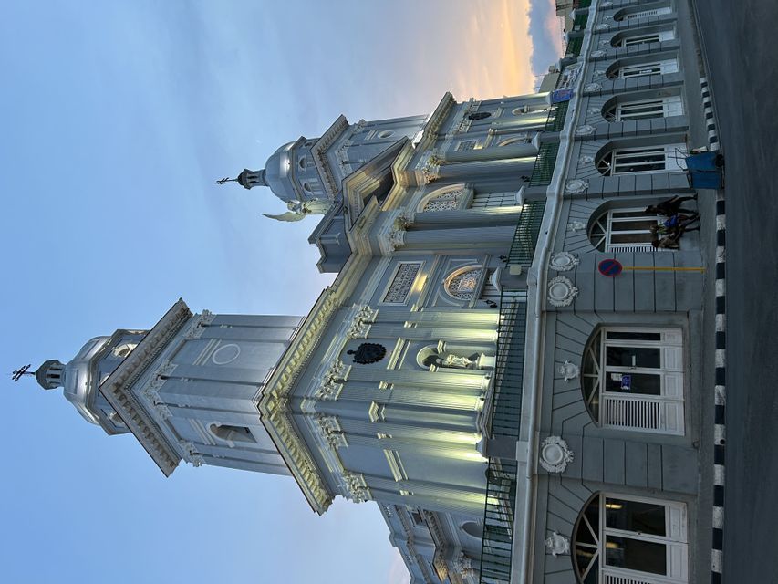 Una iglesia gris y ornamentada con dos campanarios iluminados se alza contra el cielo al anochecer, con dos personas sentadas en una cornisa.