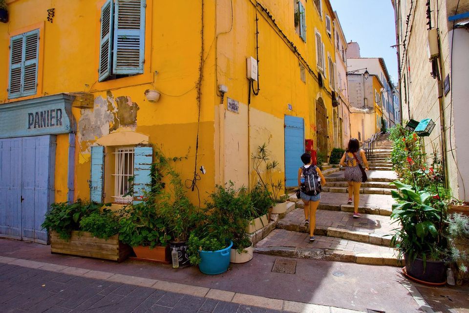 Due donne salgono una scalinata di pietra in un vicolo stretto fiancheggiato da edifici gialli, persiane blu e piante in vaso.