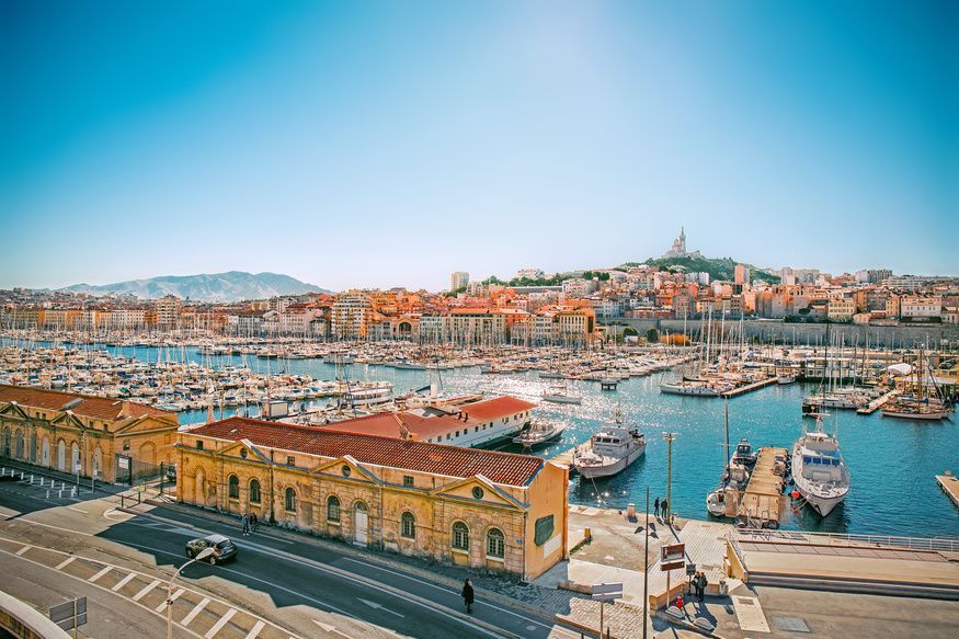 Una vista elevada y soleada de un gran puerto lleno de barcos, con una ciudad extensa y una basílica en la cima de una colina al fondo.
