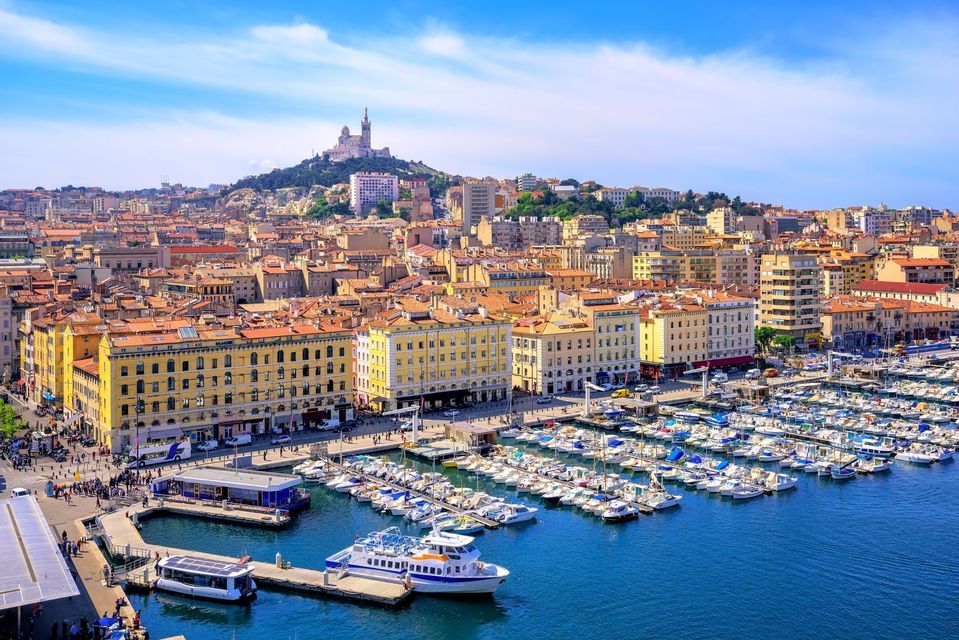 Una ciudad densa con tejados de terracota rodea un puerto lleno de barcos, con una gran basílica visible en una colina distante bajo un cielo azul.