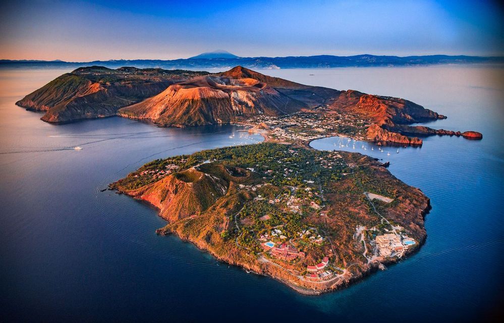 An aerial view of a volcanic island with a large crater and a town on the coast, bathed in the warm light of sunset.