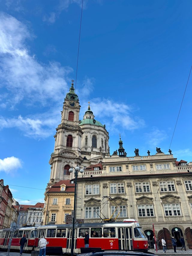Un tramway rouge et blanc passe devant des bâtiments historiques européens, avec une grande église dotée d'un dôme vert sur un ciel bleu.