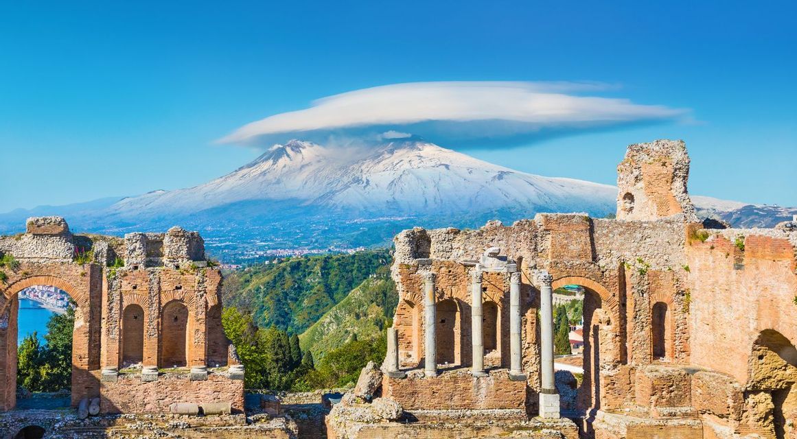 Ancient brick theater ruins frame a distant, snow-capped volcano under a clear blue sky with a single cloud.