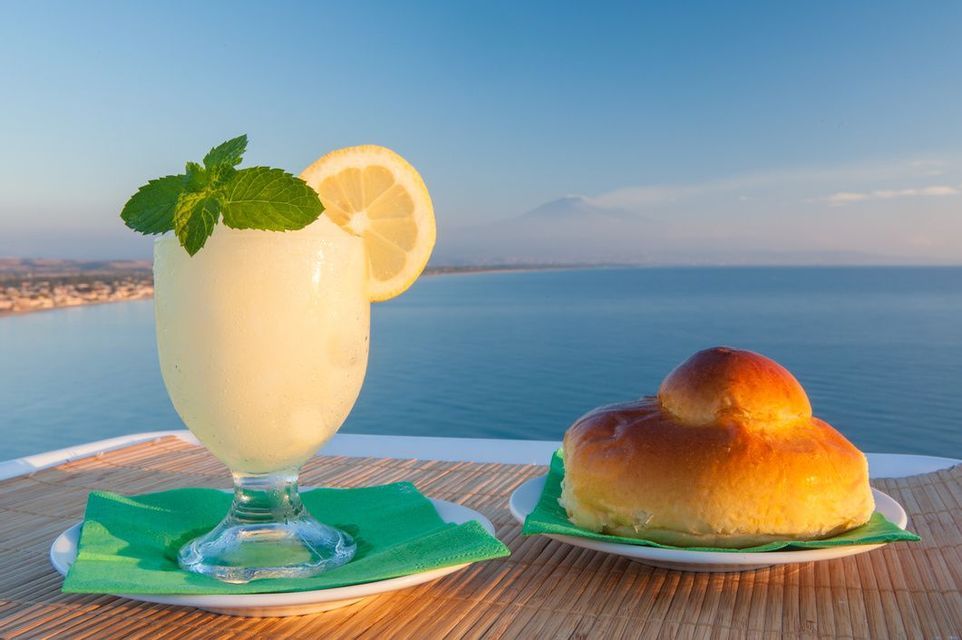 Un bicchiere di granita al limone con guarnizione di menta e una brioche su un tavolo con vista sul mare e un vulcano.