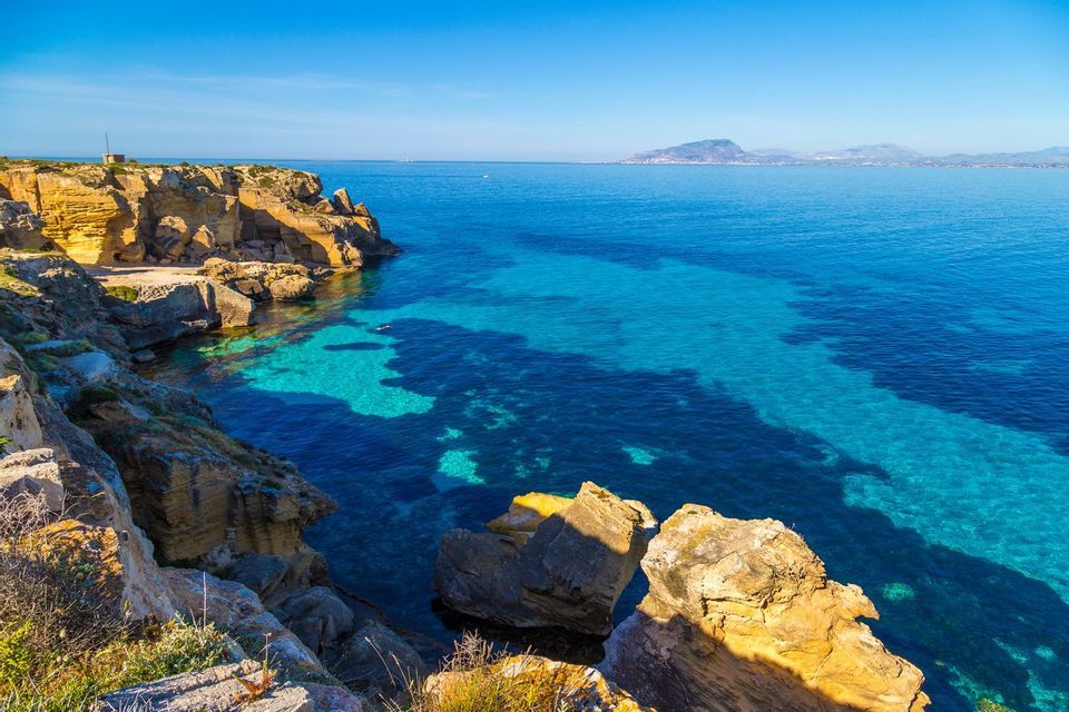 Una costa rocciosa con scogliere gialle si affaccia su un mare calmo, trasparente e turchese sotto un cielo azzurro limpido, con un'isola lontana all'orizzonte.