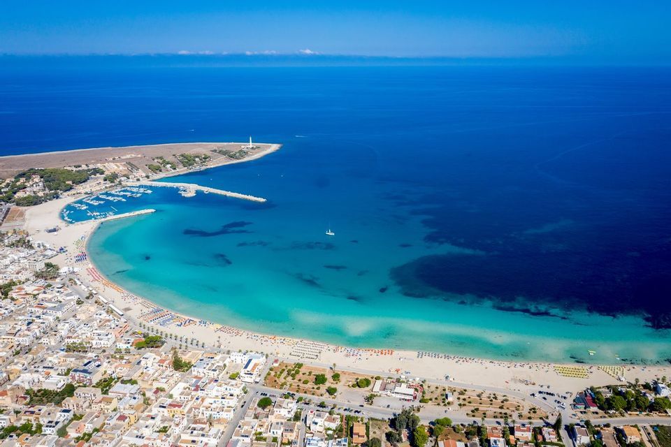Vista aerea di una città costiera con una spiaggia a mezzaluna, un porto turistico con una diga foranea e un faro su una penisola.