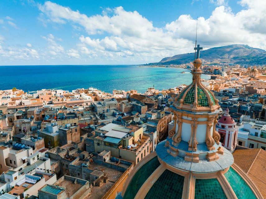 Vista aerea di una cupola di chiesa con tegole verdi che si affaccia su una città costiera con acqua azzurra e montagne sotto un cielo parzialmente nuvoloso.