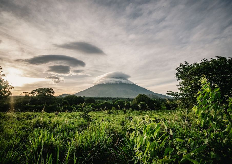Un volcan avec un nuage lenticulaire au-dessus de son sommet, s'élevant au-dessus d'un paysage verdoyant de champs et de forêt au coucher du soleil.