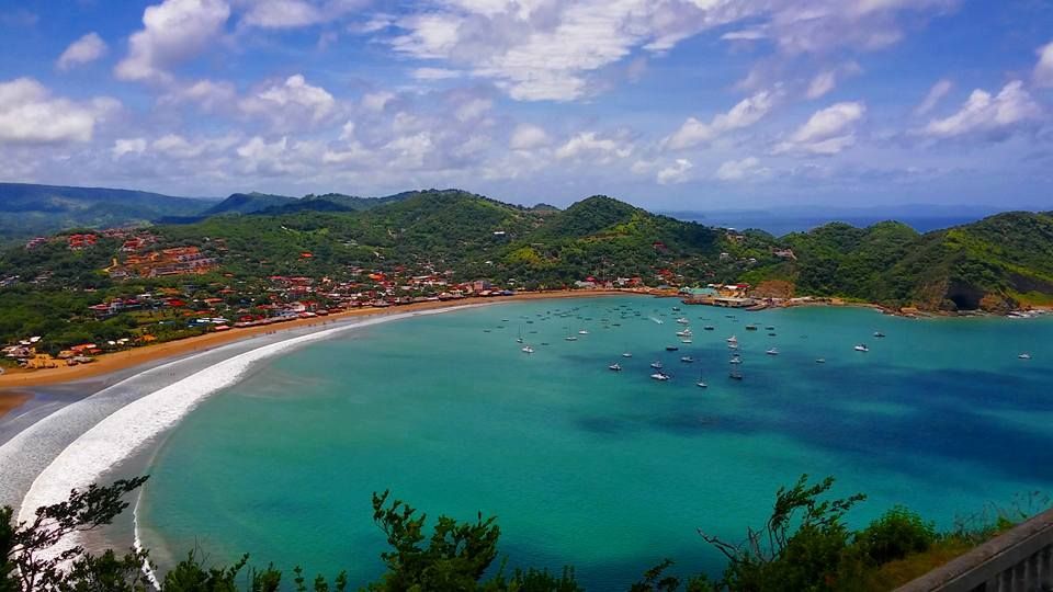 Vue plongeante d'une baie turquoise avec de nombreux bateaux ancrés près d'une plage de sable et d'une ville côtière entourée de collines verdoyantes.