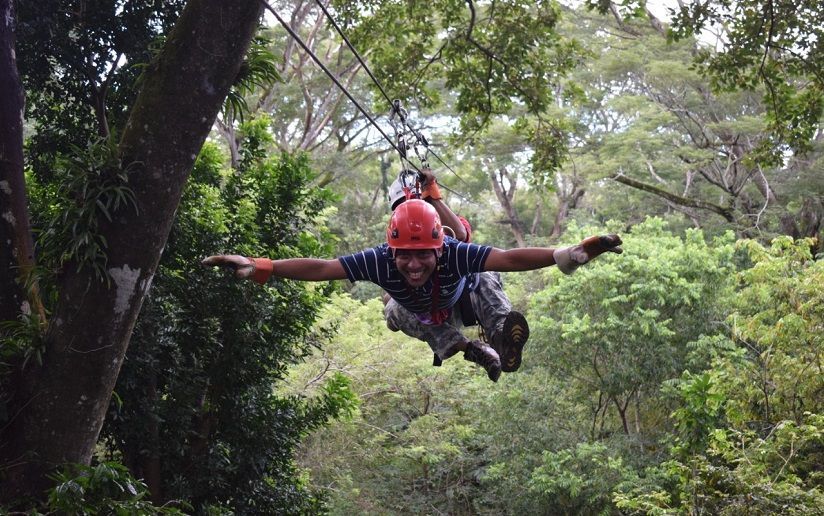 Un homme souriant en casque rouge fait de la tyrolienne à travers une forêt verte, les bras grands ouverts.