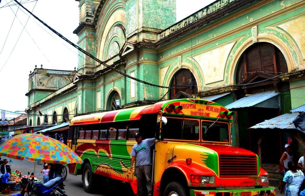 Un bus aux couleurs vives rouge, jaune et vert garé dans une rue de ville devant un grand bâtiment de style colonial vert.
