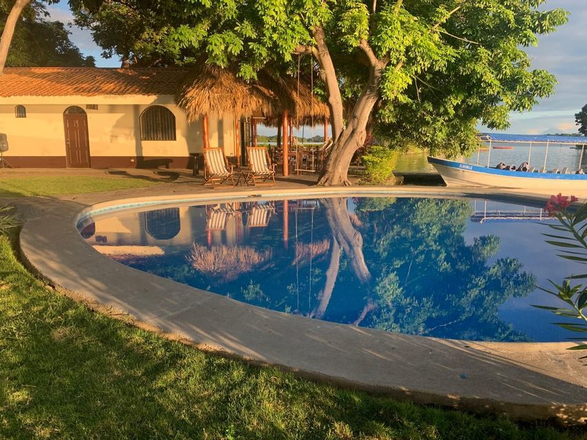 Une piscine incurvée reflète un grand arbre et un bâtiment à l'heure dorée, avec un bateau sur un lac en arrière-plan.