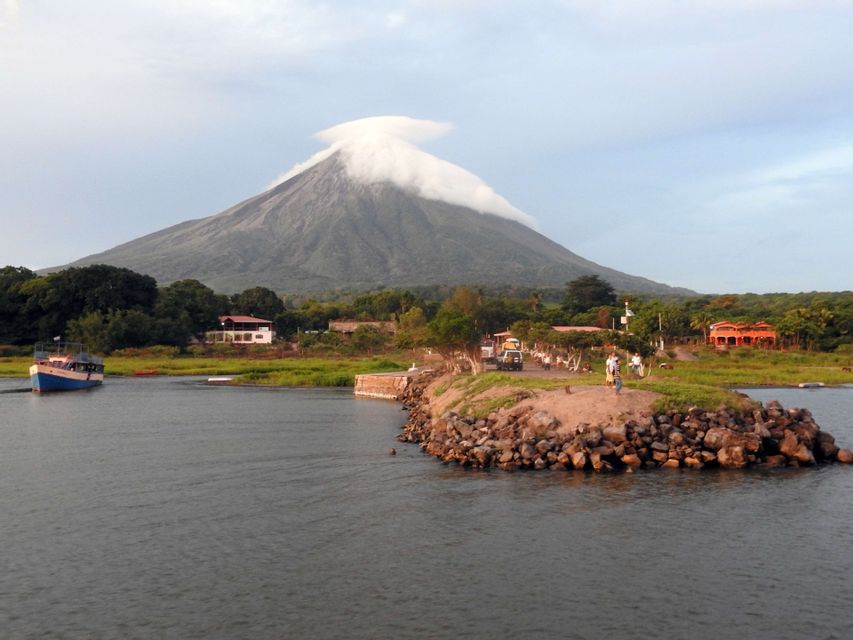 Un volcan avec un nuage lenticulaire sur son sommet surplombe un plan d'eau avec un bateau et une jetée rocheuse au premier plan.