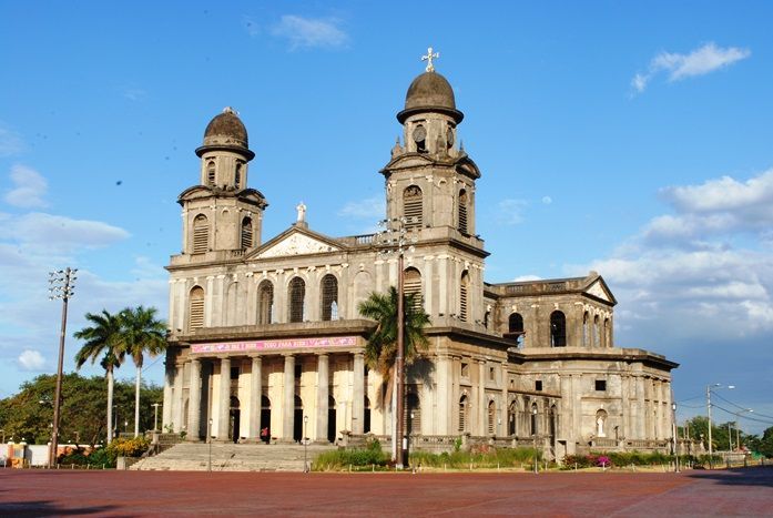 Une grande cathédrale de pierre avec deux clochers à dôme et une entrée à colonnes, vue depuis une place en briques rouges.