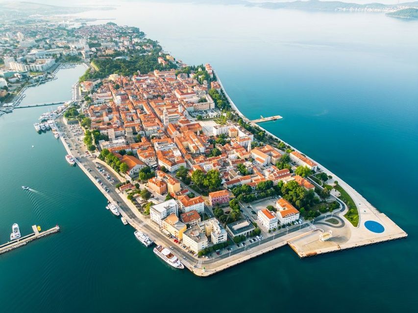 Una vista aerea di una città costiera su una penisola, con edifici dai tetti in terracotta circondati dal mare.