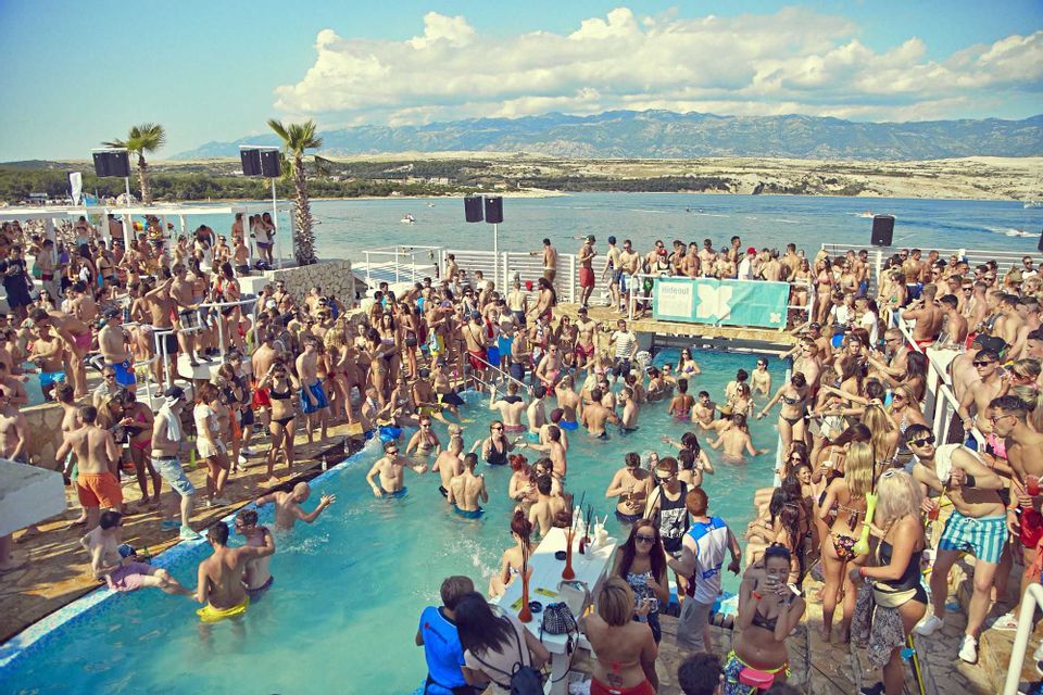 Un viaggio di gruppo WeRoad a una festa in piscina in un affollato beach club, con il mare e le montagne distanti sullo sfondo.