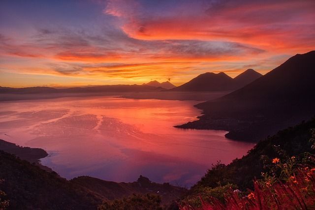 Un tramonto colorato con nuvole arancioni e viola si riflette su un grande lago circondato da montagne e vulcani in silhouette.