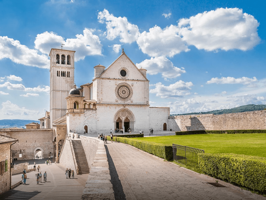 Una grande basilica storica in pietra con un campanile e un rosone, vista da un sentiero di pietra in una giornata di sole con nuvole.
