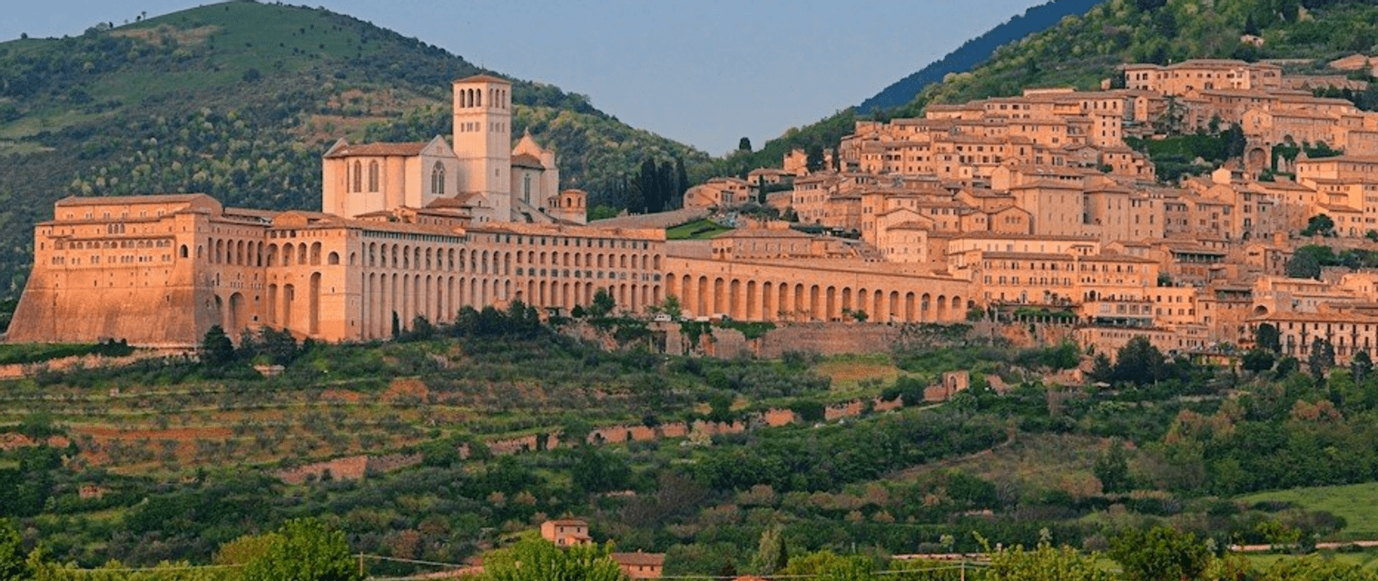 Vista panoramica di una città storica e di una basilica incastonate in una verdeggiante collina al tramonto.
