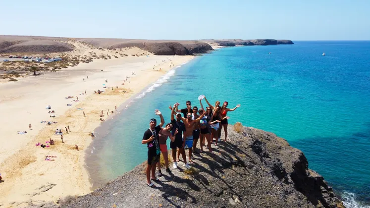 Una veduta aerea di un gruppo WeRoad che saluta da una scogliera rocciosa a picco su una lunga spiaggia sabbiosa e acqua turchese.