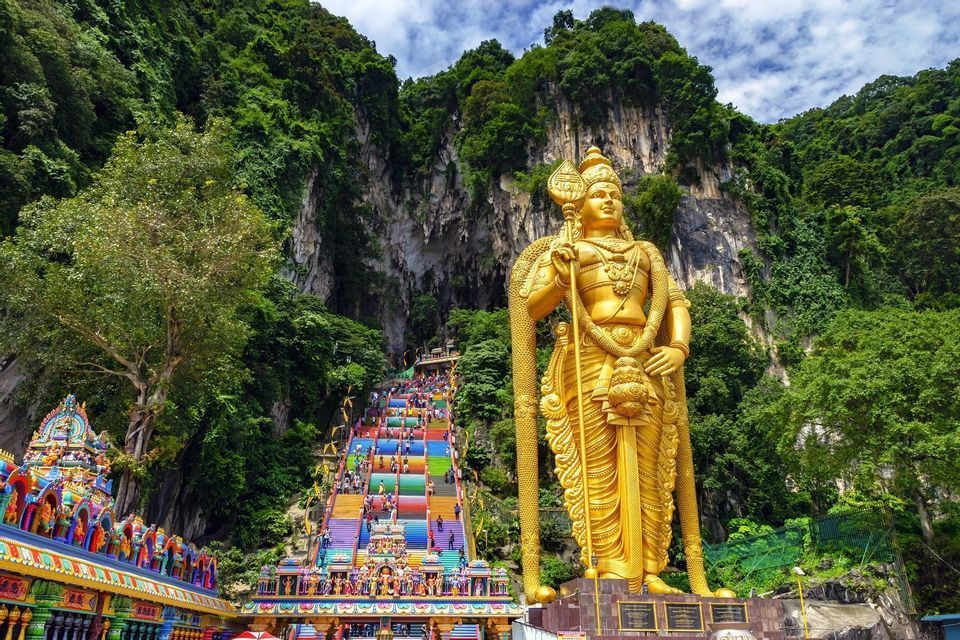 Una gigantesca statua dorata si erge accanto a una lunga scalinata dai colori dell'arcobaleno che porta su una rigogliosa scogliera verde, mentre le persone la salgono.