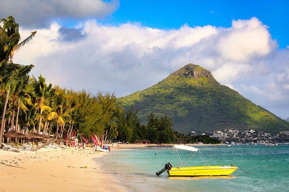 Un bateau à moteur jaune repose dans l'eau turquoise d'une plage de sable bordée de palmiers, avec une grande montagne verte en arrière-plan.