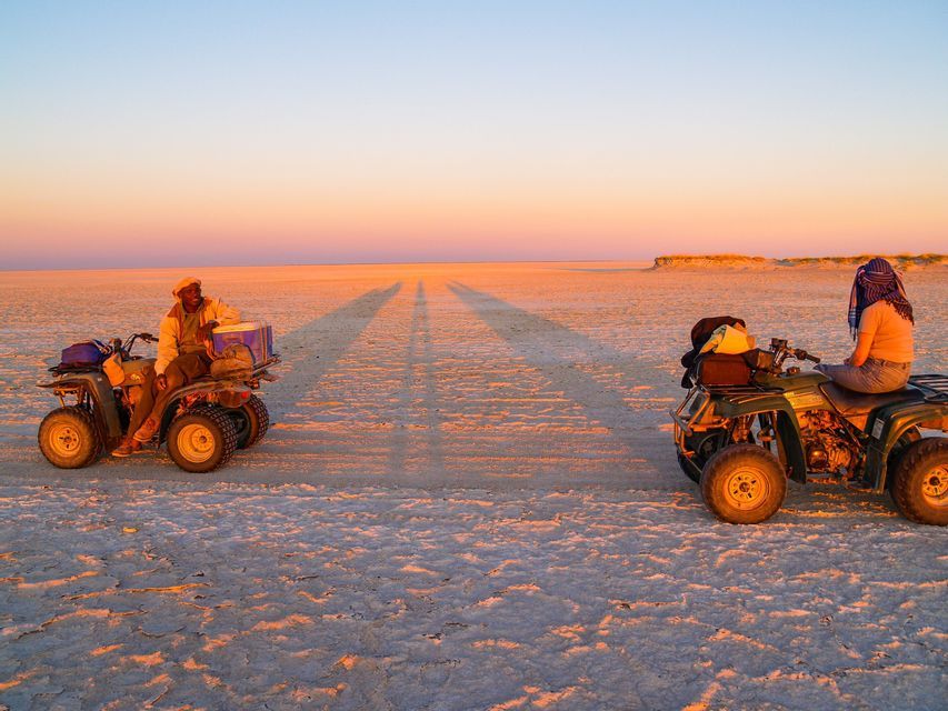 Due partecipanti a un viaggio di gruppo WeRoad sono fermi sui quad in una vasta distesa salata al tramonto, proiettando lunghe ombre.