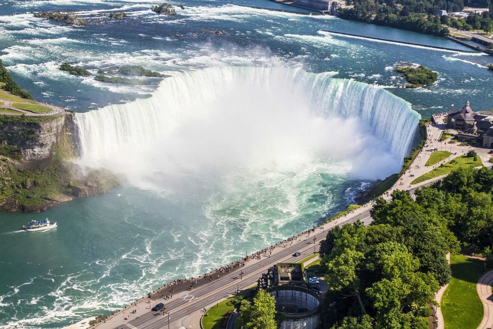 Una vista aerea di una massiccia cascata a forma di ferro di cavallo che si getta in un fiume, con una barca turistica sotto e persone su una passeggiata vicina.