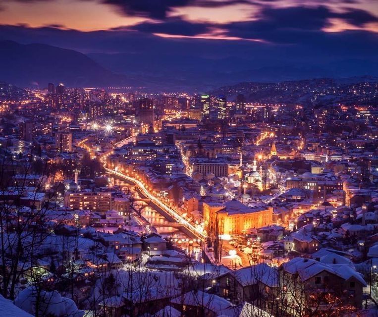 Un paisaje urbano al anochecer con edificios nevados y farolas brillantes, visto desde un punto elevado.
