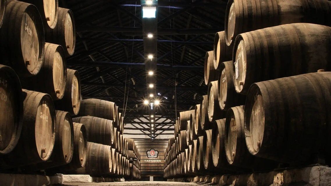 Hileras de grandes barricas de madera están apiladas en lo alto en una bodega tenuemente iluminada con un techo alto de vigas y un suelo de piedra.