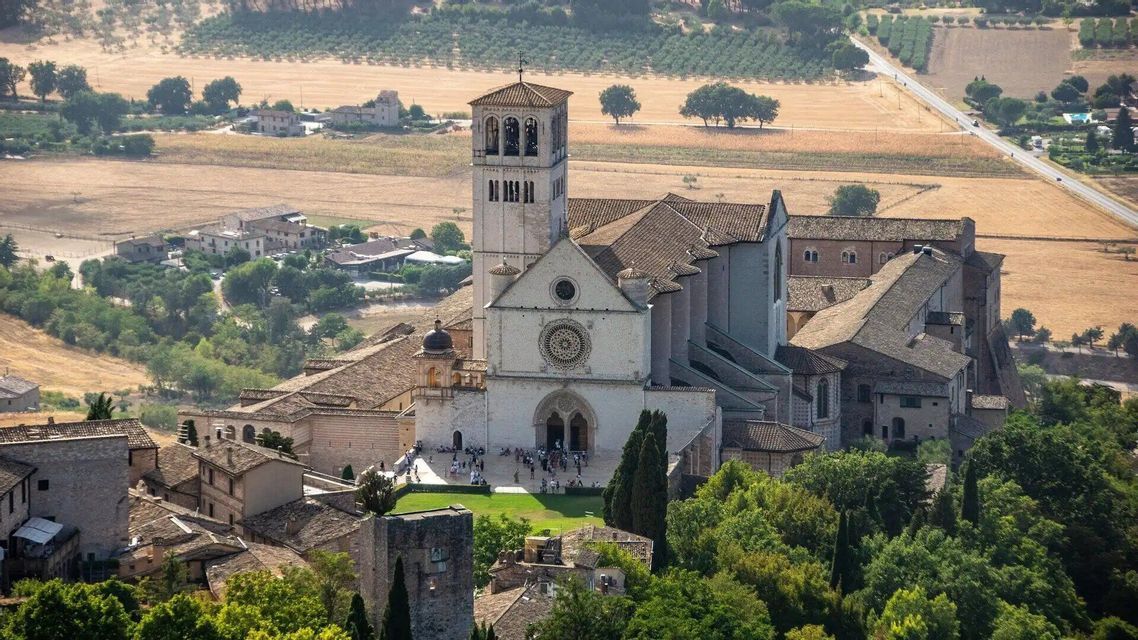 Vista aérea de una gran basílica de piedra con un campanario en una ladera, dominando un valle con campos y árboles.