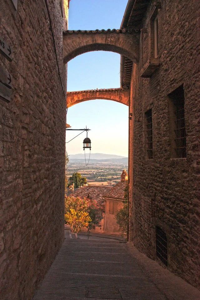 Arcos de piedra enmarcan una vista por un callejón escalonado, con vistas a un valle soleado con tejados de terracota y colinas.