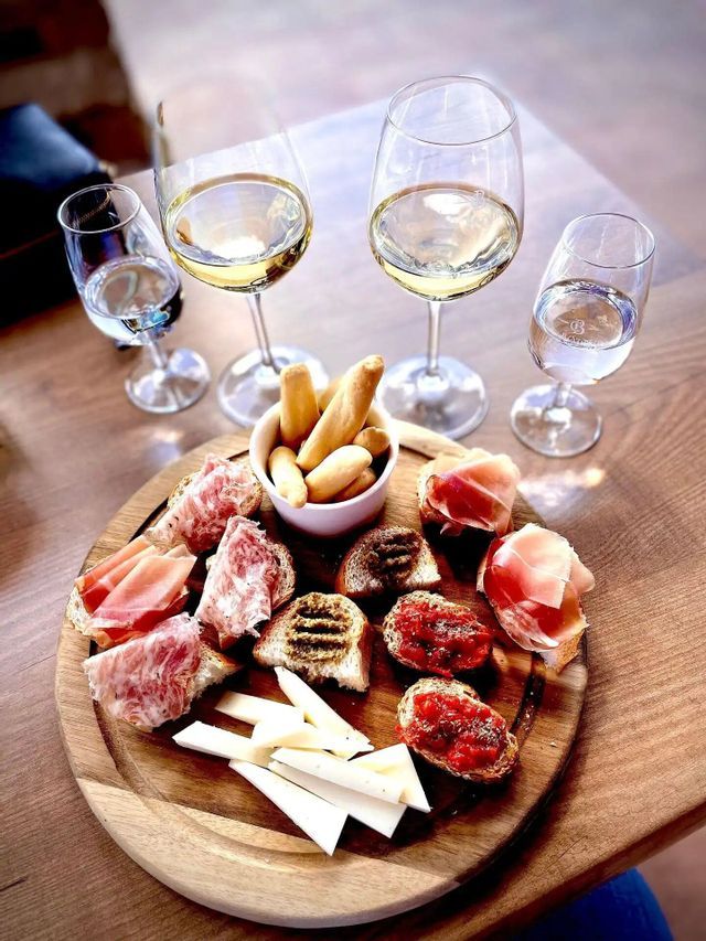 Una tabla de madera contiene aperitivos como crostini, embutidos y queso, servidos junto a copas de vino blanco y agua.